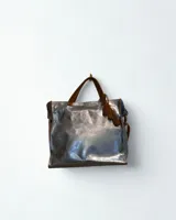 Bolso tipo tote bag plateado metalizado con doble asa corta marrón, correa larga ajustable y detalle de flecos de cuero marrón. Interior forrado en color azul.