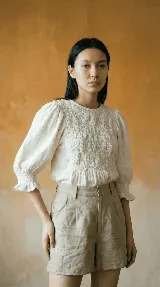 Blusa de color natural confeccionada en tejido 100% algodón. Presenta un cuerpo delantero fruncido tipo smock, vuelo peplum en la cintura y mangas globo 3/4 con volados en los puños.