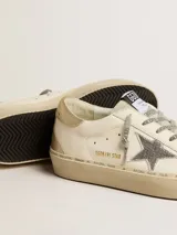 Championes de cuero Golden Goose modelo Hi Star, color blanco con detalles en dorado y plateado. Presentan una estrella de glitter plateado en el lateral, puntera de gamuza gris y talón en cuero dorado. La suela de plataforma incluye inscripciones de la marca y un acabado sutilmente desgastado.
