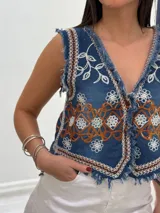 Chaleco corto de denim azul con bordados florales en blanco y marrón, detalle desflecado en escote, sisas y ruedo. Escote en V y cierre frontal con botones. Estilo boho chic.