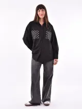 Camisa negra de corte recto, con cuello clásico, cierre frontal con botones y mangas largas con puños abotonados. Presenta dos bolsillos de parche en el pecho con apliques de tachas blancas formando un patrón geométrico.