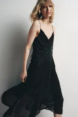 Vestido midi negro de tirantes finos con escote fluido y detalle de abalorios en el corpiño. Falda asimétrica con volados. Forro interior.