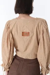Camisa color beige con textura, escote en V, mangas largas abullonadas con lazos en los puños y cierre frontal con botones.