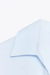 Camisa celeste de popelín con cuello solapa, escote pico y manga corta. Presenta dos bolsillos delanteros de parche y cierre frontal con botones.