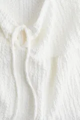 Top corto blanco de punto arrugado con escote corazón, manga circular, sección cut-out y cordón para ajustar en el busto. Modelo ajustado sobre el busto con copas preformadas fruncidas y peplum con leve vuelo.