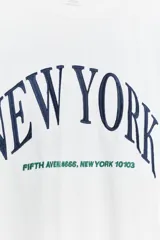Camiseta blanca de algodón con cuello redondo, corte oversize y mangas cortas. Estampado frontal con la frase "New York" en azul.