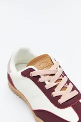 Zapatilla urbana de mujer con silueta retro, combinando paneles de gamuza marrón oscuro y material color crema en el cuerpo. Presenta detalles en color bordó en el talón y cordones beige. La suela es robusta de color marrón claro con relieve.