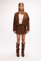 Chaqueta de efecto gamuza en color tostado, con solapas amplias, cierre frontal con un botón y flecos largos decorativos en la parte frontal y trasera de las mangas. Presenta una estructura definida y forro interior.