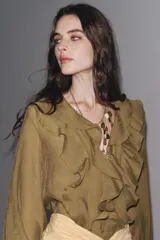 Camisa color verde oliva de tela traslúcida, con escote en V y volantes en el frente y cuello. Mangas largas abullonadas con puños elásticos.