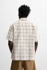 Camisa de manga corta color crudo con estampado de cuadros en tonos grises y marrones. Tiene cuello bowling, corte relaxed fit, aberturas laterales en el bajo y cierre frontal con botones.
