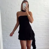 Vestido negro, sin mangas, con escote asimétrico y detalle de lazo o corbatín largo que cae desde el hombro. El cuerpo es ajustado y fruncido, y la falda corta tiene volados superpuestos.
