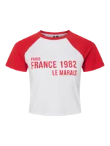 Camiseta blanca con mangas cortas raglán rojas y estampado frontal con la leyenda "Paris France 1982 Le Marais" en rojo.