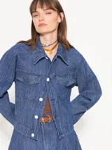 Campera de denim azul con textura en relieve, corte recto, cuello camisero, cierre frontal con botones blancos, bolsillos delanteros y mangas largas con pinzas que generan volumen.