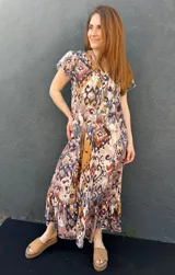 Vestido largo con estampado abstracto en tonos marrones, azules y beige, con escote en V, mangas cortas con volados y falda con volados en el ruedo.