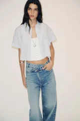 Camisa blanca corta con rayas verticales celestes, cuello solapa y mangas cortas con dobladillo. Tiene un bolsillo de parche en el pecho y cierre frontal con botones ocultos.