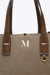 Bolso tipo shopper color marrón con doble asa corta y correa larga extraíble. Tiene detalle de pespuntes y anilla metálica decorativa. Incluye bolsa de tela interior extraíble con cierre fruncido.
