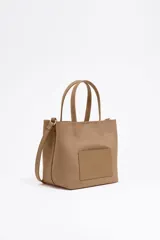 Bolso tote pequeño color marrón claro, de la marca Bimba y Lola, modelo Chihuahua. Tiene doble asa corta y correa larga extraíble. Presenta un bolsillo externo con parche con el logo de la marca.