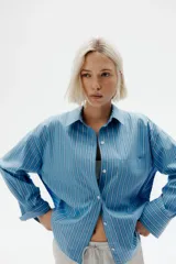 Camisa celeste y blanca a rayas verticales, de corte amplio y mangas largas. Tiene cuello clásico, cierre frontal con botones y bolsillo de parche en el pecho.
