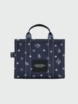Bolso tipo tote de lona color azul marino con diseño gráfico estampado en color blanco. Incluye el nombre de la marca 'Marc Jacobs' y el modelo 'The Tote Bag' en el frente, con detalles de estrellas, corazones y flores. Posee doble asa superior, correa de hombro ajustable y costuras en contraste.