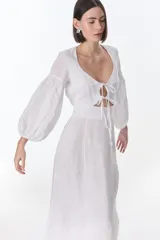 Vestido color beige de lino, con escote en V, mangas largas abullonadas y lazos frontales. Tiene corte a la cintura y falda larga con tajo frontal.