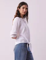 Blusa blanca de tejido texturado con cuello redondo, escote en V con aplique metálico y mangas 3/4 con puños elásticos. El ruedo se ata con un nudo.