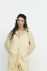Camisa oversize a rayas amarillas y blancas, de cuello solapa y manga larga tipo murciélago acabada en puño con hombro caído. Tiene un bolsillo de plastrón en el delantero y cierre frontal con botones.