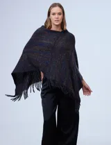 Poncho tejido en hilo soft, con efecto jaspeado multicolor (predominantemente tonos oscuros como azul marino, negro, y toques de violeta y dorado/lurex). Presenta un diseño asimétrico y flecos largos en los bordes.