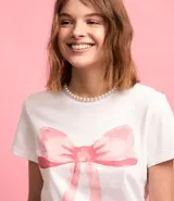 Remera blanca de algodón con cuello redondo y mangas cortas. Presenta una estampa de una moña rosa en el frente.