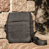 Morral de tela canvas color verde oliva, con tapa, apertura superior y cierre. Tiene bolsillos en la tapa al frente, uno con cierre, y un bolsillo auxiliar también al frente con cierre bajo la tapa. Incluye un bolsillo con cierre atrás y correa larga regulable.