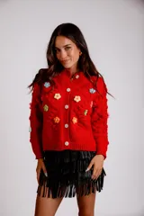 Saco de punto color rojo, con cuello polo y cierre frontal de botones blancos. Presenta flores bordadas en relieve de diferentes colores (azul, amarillo, verde, naranja, rosado) y mangas largas con diseño de burbujas.