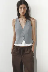 Chaleco gris con rayas verticales, escote en pico y cierre frontal con botones. Detalle de camisa blanca en el bajo.