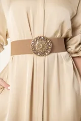 Cinto de malla elástica color beige, con hebilla metálica circular adornada con piedras y strass.