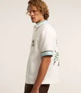 Camisa masculina de algodón con corte boxy, cuello con solapa y rayas, abotonado delantero, bolsillo con estampa floral y estampa trasera de palmera con la palabra 'Paradise'.