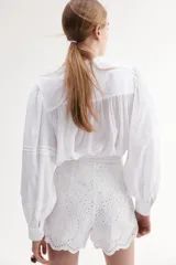 Blusa blanca de algodón con cuello mao, pechera con jaretas e inserciones de encaje, botones frontales, canesú fruncido en la espalda y mangas largas abullonadas con pinzas y abertura con botón en los puños.