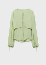 Blusa color verde claro de gasa semitransparente con cuello redondo y lazos en el frente. Tiene mangas largas abullonadas con puños elásticos y fruncidos. Incluye un chaleco corto incorporado. 