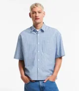 Camisa masculina oversized boxy de manga corta, confeccionada en tejido oxford a rayas verticales celestes y blancas. Cuenta con cuello inglés, abotonadura frontal y bolsillo de parche en el pecho.