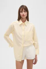 Camisa holgada de algodón color amarillo pálido con rayas verticales. Tiene cuello camisero, manga larga con puños abotonados y un bolsillo de parche en el pecho.