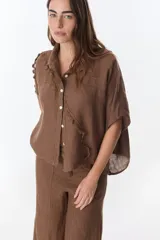 Camisa color beige de lino con cuello camisero, manga corta y cierre frontal con botones. Presenta detalles de encaje en el cuello, el frente y las mangas.