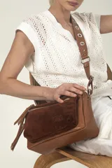 Bandolera de cuero color marrón con bolsillo frontal de gamuza color óxido. Tiene correa regulable con ojalillos metálicos, cierre superior y herrajes metálicos.