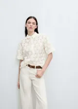 Blusa color crema con textura calada de flores en relieve, cuello camisero, manga corta y cierre de botones en el cuello.