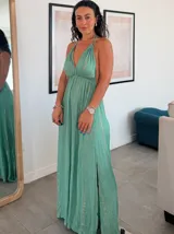 Vestido largo blanco con finas rayas verticales en lurex dorado. Presenta un escote profundo en V con tirantes finos que se anudan en la nuca (estilo halter) y un corte imperio con frunces en la cintura.