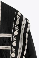 Blusa negra de algodón con cuello redondo y manga larga abullonada. Presenta bordados blancos a contraste en el cuello, hombros y mangas. Cierre en la espalda con abertura y botón.