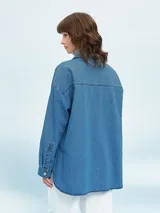 Camisa de denim negro con corte oversize, cuello clásico, cierre frontal con botones metálicos y bolsillo de parche en el pecho.