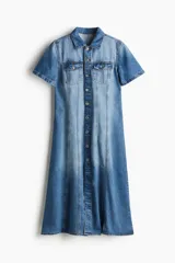 Vestido midi de denim celeste, con cuello camisero, manga corta y botonadura frontal. Presenta bolsillos superiores con solapa y botón, canesú delantero y trasero, falda acampanada y costuras decorativas.