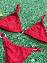 Bikini de dos piezas color rojo, con corpiño triángulo que tiene una argolla central de zamak y bombacha colaless con argollas laterales. La tela es texturada.