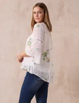 Blusa blanca de gasa con bordados florales, corte holgado, mangas 3/4 acampanadas con detalle de puntilla y forro interno.