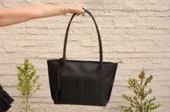 Cartera tote de cuero vacuno color negro, con forrería y cierre. Presenta un bolsillo externo con detalle de costuras y flecos.