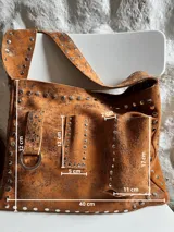 Cartera bandolera marrón de cuero con remaches metálicos plateados. Tiene correa ancha regulable, bolsillo externo y argolla para colgar accesorios.