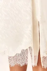 Vestido lencero midi color blanco roto, con tirantes finos y escote en V adornado con encaje. El tejido presenta un patrón sutil en relieve y el ruedo termina con un borde de encaje.