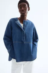 Blusa azul de denim con cuello redondo, tapeta con botones ocultos y mangas largas con puños abotonados.